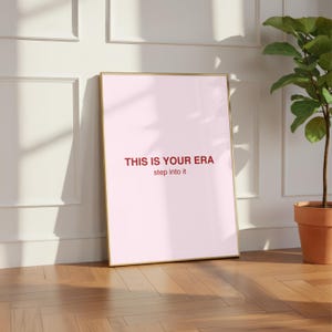 Könnte beinhalten: Ein gerahmter rosa Kunstdruck mit dem Text "THIS IS YOUR ERA step into it" in roter Schrift. Der goldene Rahmen lehnt an einer weißen Wand und einem Holzboden. Eine Topfpflanze befindet sich im Hintergrund.