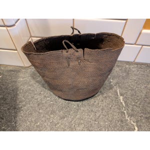 Può includere: Un cesto marrone intrecciato con una parte superiore ampia e aperta e una base arrotondata. Il cesto ha due manici ed è realizzato in un materiale strutturato. Il cesto è su una superficie grigia.