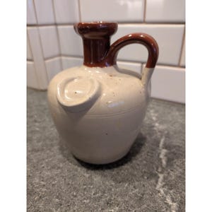 Pode incluir: Jarro de cerâmica branco sujo com alça e bico marrons. O jarro tem um corpo arredondado e um desenho circular em relevo na frente. A alça e o bico são de cor marrom escuro. O jarro é provavelmente vintage ou antigo.