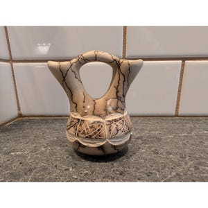 Puede incluir: Un jarrón de cerámica beige con un asa única y intrincados patrones de vetas negras. El jarrón presenta un diseño tallado alrededor del centro y se exhibe sobre una superficie gris moteada.