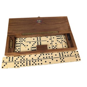 Könnte beinhalten: Ein Dominospiel aus Holz in einer rechteckigen Schachtel mit Klappdeckel. Die Schachtel hat ein messingfarbenes Metallscharnier und ein kleines rundes Etikett. Im Inneren befinden sich cremefarbene Dominosteine mit schwarzen Punkten.