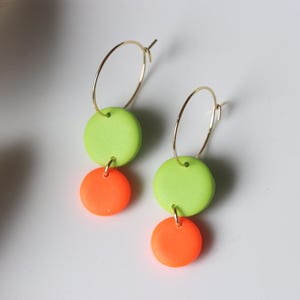 Könnte beinhalten: Ein Paar goldene Creolen mit einem Design aus neongrünen und orangefarbenen Scheiben. Die Ohrringe haben einen goldenen Reifen mit einer grünen und einer orangefarbenen Scheibe. Ein farbenfrohes Accessoire.