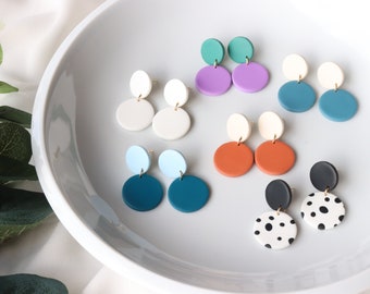 May include: A set of 10 pairs of colorful, handmade polymer clay earrings. Each pair features two round discs, one on top of the other. The colors include white, teal, light blue, purple, orange, black, and white with black polka dots.