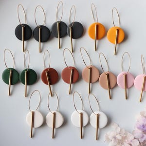 Puede incluir: Un conjunto de 12 pares de pendientes de aro dorados con una barra dorada y un disco redondo de arcilla en varios colores. Los colores incluyen negro, verde, marrón, naranja, beige, rosa y blanco.