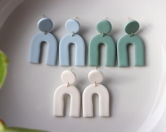 May include: Three pairs of handmade polymer clay earrings. The earrings are in the shape of an arch with a round disc at the top. The colors are light blue, teal, and white.