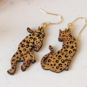 Könnte beinhalten: Ein Paar Holz-Ohrringe mit Leopardenmuster. Ein Ohrring zeigt einen springenden Leoparden, der andere einen sitzenden. Beide Ohrringe haben goldfarbene Ohrhaken und ein geflecktes Muster.