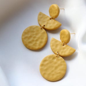 Könnte beinhalten: Senfgelbe Ohrringe mit strukturierter Oberfläche. Jeder Ohrring hat ein kreisförmiges und ein halbkreisförmiges Element, verbunden durch eine kleine goldfarbene Schlaufe. Auf weißem, seidigem Hintergrund.