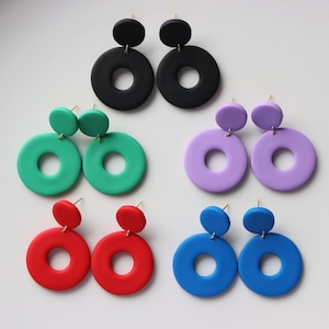 Könnte beinhalten: Eine Sammlung farbenfroher, handgefertigter Ohrringe in verschiedenen Farbtönen. Die Ohrringe haben ein kreisförmiges Design mit einem kleineren Kreis oben. Farben sind Schwarz, Grün, Lila, Rot und Blau. Die Ohrringe werden auf einer weißen Oberfläche präsentiert.