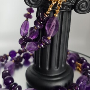 May include: A purple amethyst gemstone necklace with gold beads and a gold clasp. The necklace is displayed on a black column.