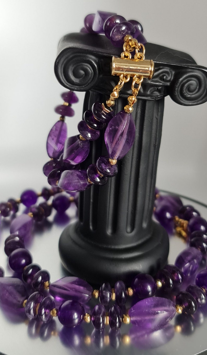 May include: A purple amethyst gemstone necklace with gold beads and a gold clasp. The necklace is displayed on a black column.