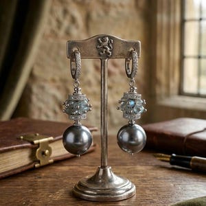May include: Silver earrings on a display stand. Each earring has a large, dark grey pearl, a cluster of blue-green stones, and a diamond-set hoop. The stand is silver and has a decorative emblem at the top.