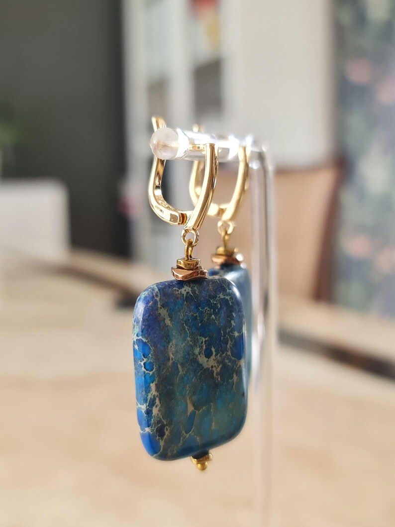 Puede incluir: Un par de pendientes colgantes de tono dorado con colgantes rectangulares de piedra veteada en azul y verde azulado. Las piedras tienen una textura natural y est&aacute;n suspendidas de cierres de palanca dorados pulidos con detalles.