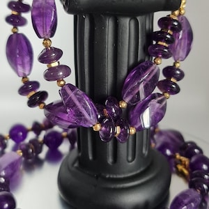 May include: A double strand necklace and bracelet set made with purple amethyst gemstone beads and gold spacers. The jewelry is displayed on a black pedestal.
