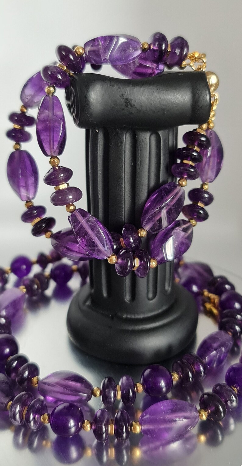 May include: A double strand necklace and bracelet set made with purple amethyst gemstone beads and gold spacers. The jewelry is displayed on a black pedestal.