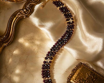 Puede incluir: Un brazalete de varias hebras con cuentas de color rojo oscuro y detalles dorados. El brazalete tiene un cierre de broche y está adornado con pequeños adornos brillantes. La joya se exhibe sobre una tela crema similar al satén.