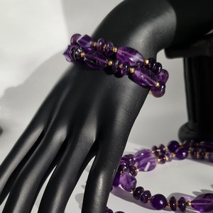 May include: A purple amethyst gemstone necklace and bracelet set with gold accents. The necklace and bracelet are made of faceted amethyst beads.