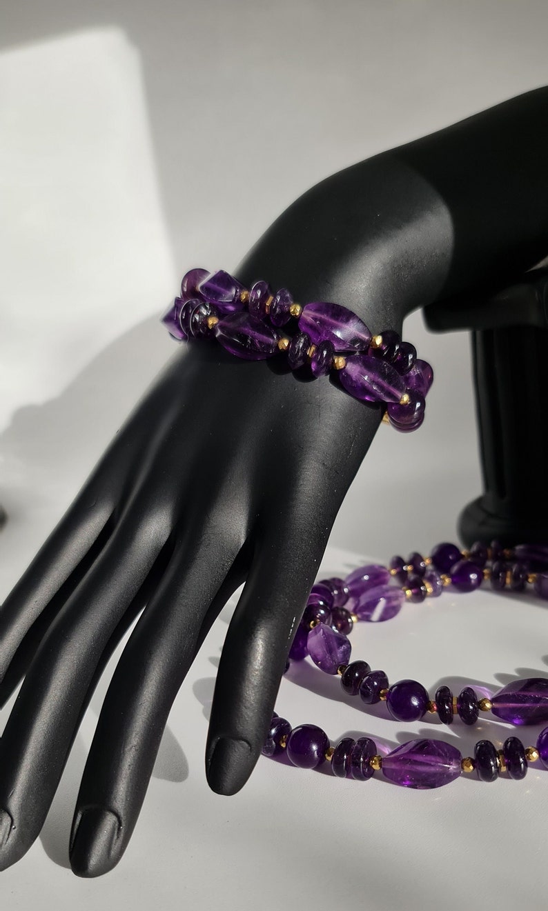 May include: A purple amethyst gemstone necklace and bracelet set with gold accents. The necklace and bracelet are made of faceted amethyst beads.