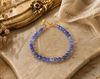 Puede incluir: Una delicada pulsera con cuentas azules facetadas ensartadas en una cadena dorada. La pulsera se exhibe sobre una superficie clara y texturizada, con una tela suave y drapeada en el fondo. Una hermosa pieza de joyería.
