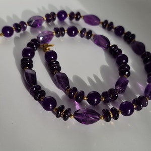 May include: A purple amethyst gemstone necklace with gold accents. The necklace is made of round and oval-shaped beads.