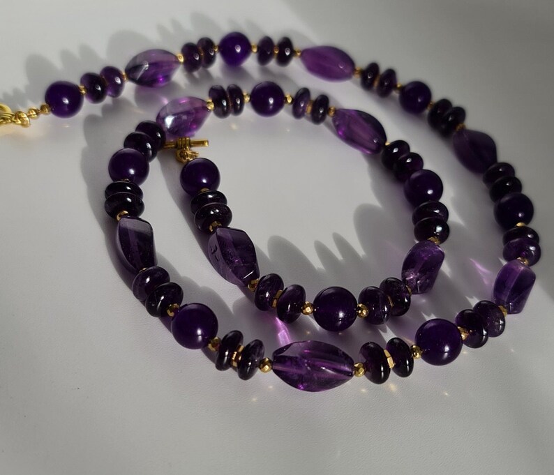 May include: A purple amethyst gemstone necklace with gold accents. The necklace is made of round and oval-shaped beads.