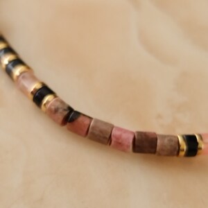 May include: A close-up of a pink and brown beaded bracelet with gold and black accents. The bracelet is made of small, square-shaped beads.