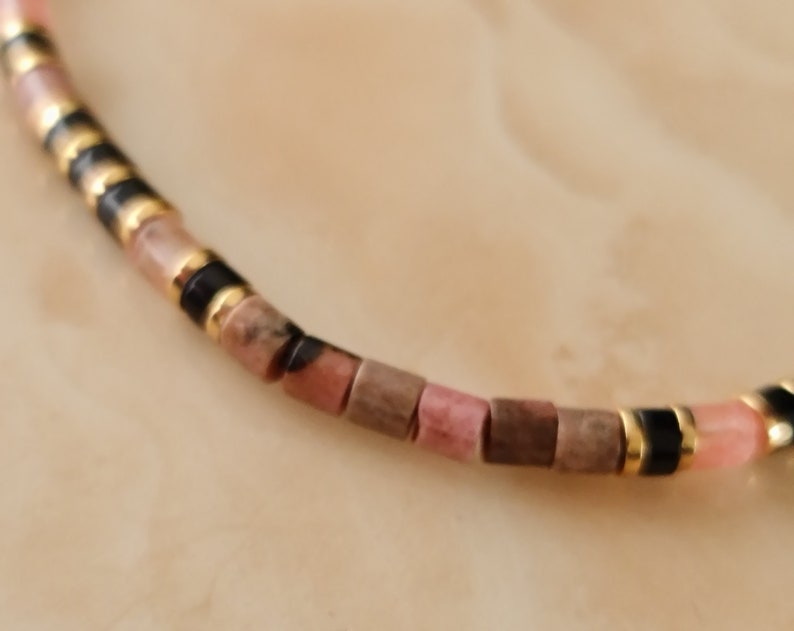 May include: A close-up of a pink and brown beaded bracelet with gold and black accents. The bracelet is made of small, square-shaped beads.