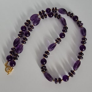 May include: A purple amethyst gemstone necklace with gold beads. The necklace has a lobster clasp closure.