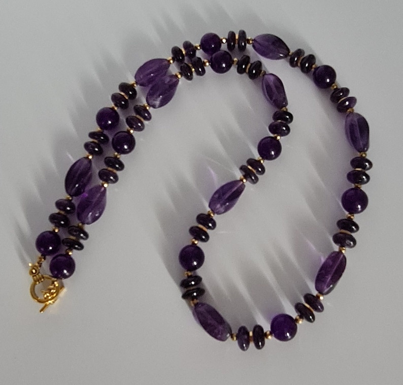 May include: A purple amethyst gemstone necklace with gold beads. The necklace has a lobster clasp closure.