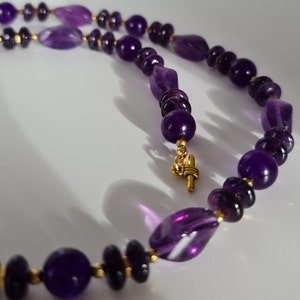 May include: A purple amethyst gemstone necklace with gold accents. The necklace features a variety of amethyst beads, including round, oval, and faceted shapes.