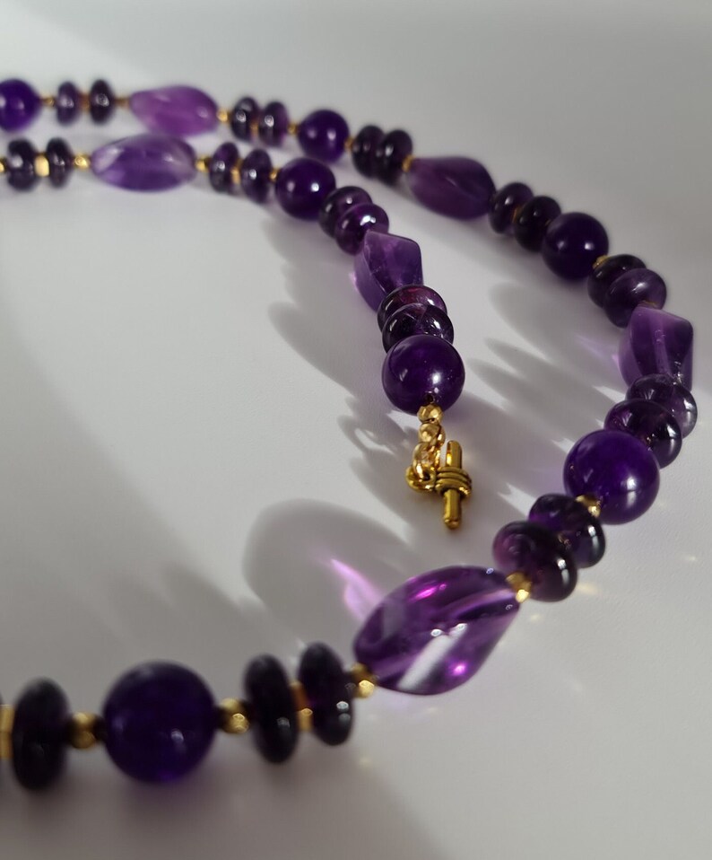 May include: A purple amethyst gemstone necklace with gold accents. The necklace features a variety of amethyst beads, including round, oval, and faceted shapes.