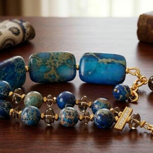 Puede incluir: Una pulsera de varias hebras con grandes cuentas rectangulares de piedra veteada azul y verde, cuentas redondas m&aacute;s peque&ntilde;as de piedra veteada azul y espaciadores de cuarzo ahumado transl&uacute;cido, con eslabones y cierre de tono dorado.