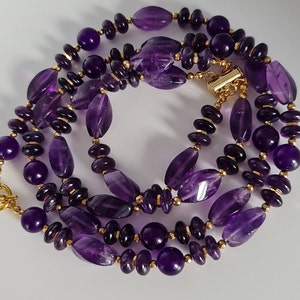 May include: A purple amethyst gemstone necklace with gold accents. The necklace is made of oval-shaped and round amethyst beads.