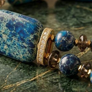 Puede incluir: Una pulsera con cuentas de piedra veteada en azul y turquesa, de formas rectangulares y esf&eacute;ricas, realzada con peque&ntilde;os espaciadores facetados transparentes y una pieza de uni&oacute;n cil&iacute;ndrica dorada con una hilera de peque&ntilde;as piedras claras.