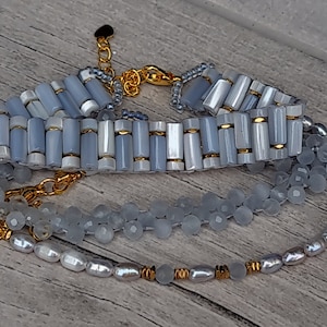 May include: Three layered necklaces with gold clasps. The top necklace is light blue with gold accents. The middle necklace is light blue with a beaded design. The bottom necklace has white pearls and gold accents.