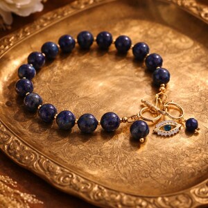 May include: A beaded bracelet featuring dark blue, round beads. The bracelet has a gold-colored clasp and a charm with an eye design. The bracelet is displayed on a gold-colored, ornate tray.