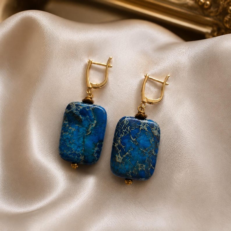 Puede incluir: Un par de pendientes colgantes con piedras rectangulares azules con intrincadas vetas oscuras. Las piedras est&aacute;n suspendidas de cierres de palanca de metal dorado, con un peque&ntilde;o abalorio dorado en la base de cada piedra rectangular.
