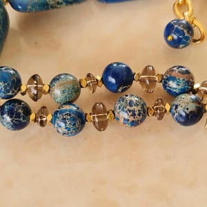Puede incluir: Una pulsera de doble hilo con cuentas redondas de jaspe jaspeado en azul y verde azulado, espaciadores de cuarzo ahumado transl&uacute;cido y peque&ntilde;os detalles met&aacute;licos dorados. La pieza termina en barras rectangulares doradas con cristales.