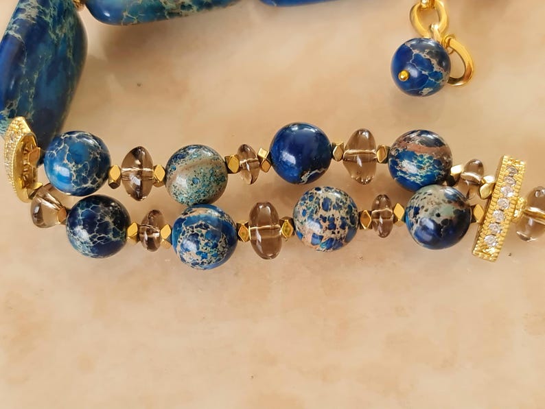 Puede incluir: Una pulsera de doble hilo con cuentas redondas de jaspe jaspeado en azul y verde azulado, espaciadores de cuarzo ahumado transl&uacute;cido y peque&ntilde;os detalles met&aacute;licos dorados. La pieza termina en barras rectangulares doradas con cristales.