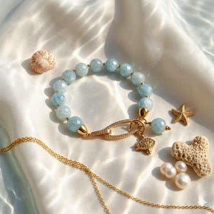 May include: A light blue beaded bracelet with gold accents and a decorative clasp. The bracelet features a small turtle charm and is surrounded by a seashell, starfish, pearls, and coral on a white satin surface.