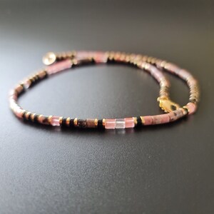 May include: A delicate pink and black beaded necklace with a gold clasp. The necklace is made with small, square-shaped beads.