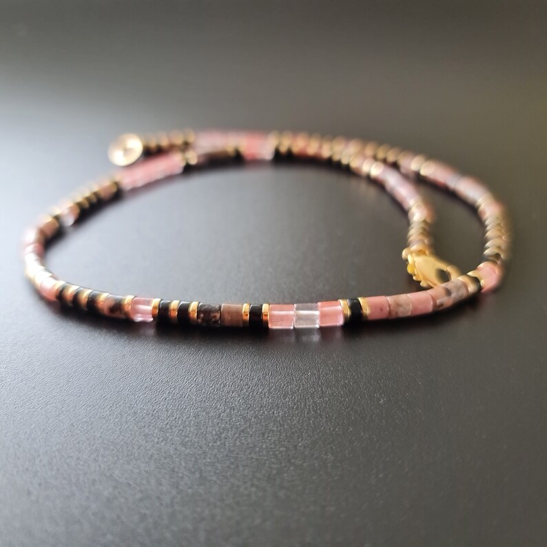 May include: A delicate pink and black beaded necklace with a gold clasp. The necklace is made with small, square-shaped beads.