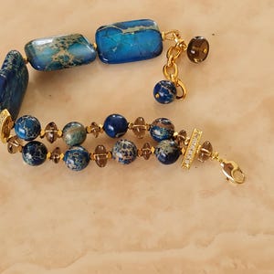 Puede incluir: Una pulsera de varias hebras con cuentas rectangulares y esf&eacute;ricas jaspeadas en azul y marr&oacute;n, intercaladas con peque&ntilde;as cuentas facetadas de color marr&oacute;n transl&uacute;cido. El dise&ntilde;o incluye detalles en metal dorado, una cadena extensora y un cierre de mosquet&oacute;n.