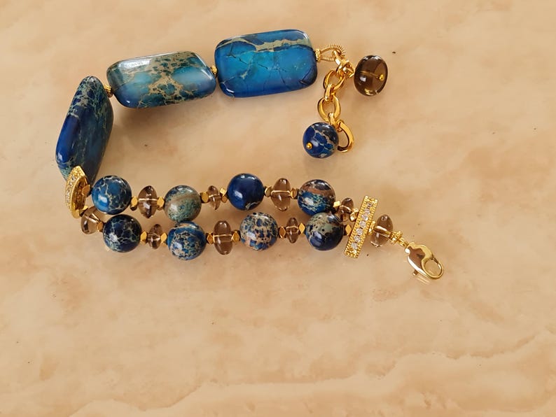 Puede incluir: Una pulsera de varias hebras con cuentas rectangulares y esf&eacute;ricas jaspeadas en azul y marr&oacute;n, intercaladas con peque&ntilde;as cuentas facetadas de color marr&oacute;n transl&uacute;cido. El dise&ntilde;o incluye detalles en metal dorado, una cadena extensora y un cierre de mosquet&oacute;n.