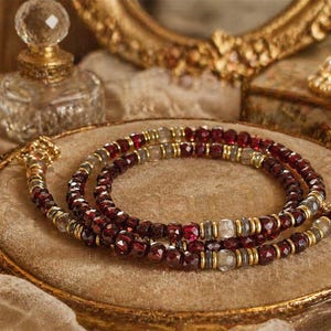 Può includere: Una collana a più strati con perline rosso scuro e trasparenti, rifinita con metallo color oro. La collana è esposta su un vassoio dorato color beige, con un flacone di profumo vintage e gioielli sullo sfondo.