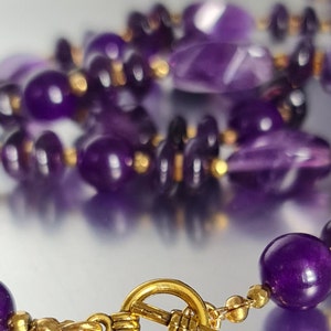 May include: A purple amethyst gemstone necklace with a gold clasp. The necklace is made of various shapes and sizes of amethyst beads.