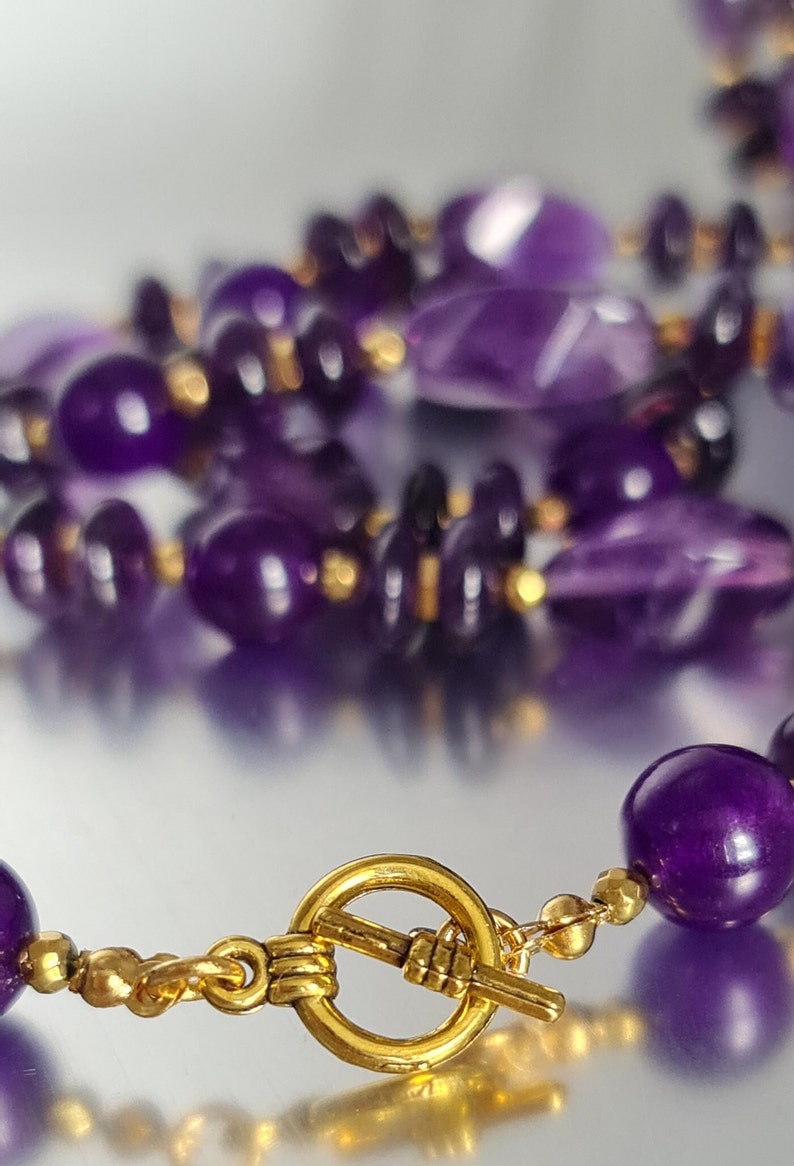 May include: A purple amethyst gemstone necklace with a gold clasp. The necklace is made of various shapes and sizes of amethyst beads.