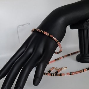 May include: A pink and black beaded necklace and matching bracelet set. The beads are small and rectangular, with a mix of pink, black, and gold accents.