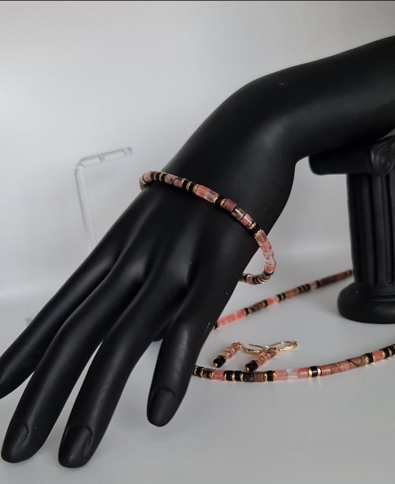 May include: A pink and black beaded necklace and matching bracelet set. The beads are small and rectangular, with a mix of pink, black, and gold accents.