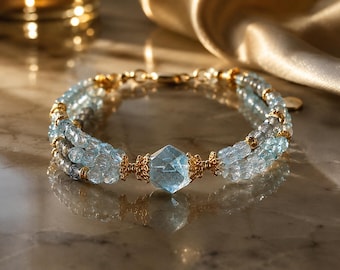 Puede incluir: Una pulsera de varias hebras con cuentas facetadas de color azul claro y una piedra preciosa hexagonal central de color azul claro. Las hebras están acentuadas con pequeños espaciadores de metal dorado y filigranas.