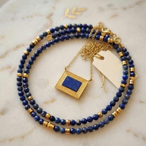 May include: A layered necklace featuring small, dark blue beads and gold-colored accents. A gold pendant with a rectangular, dark blue stone hangs from the necklace. The jewelry is displayed on a white surface.